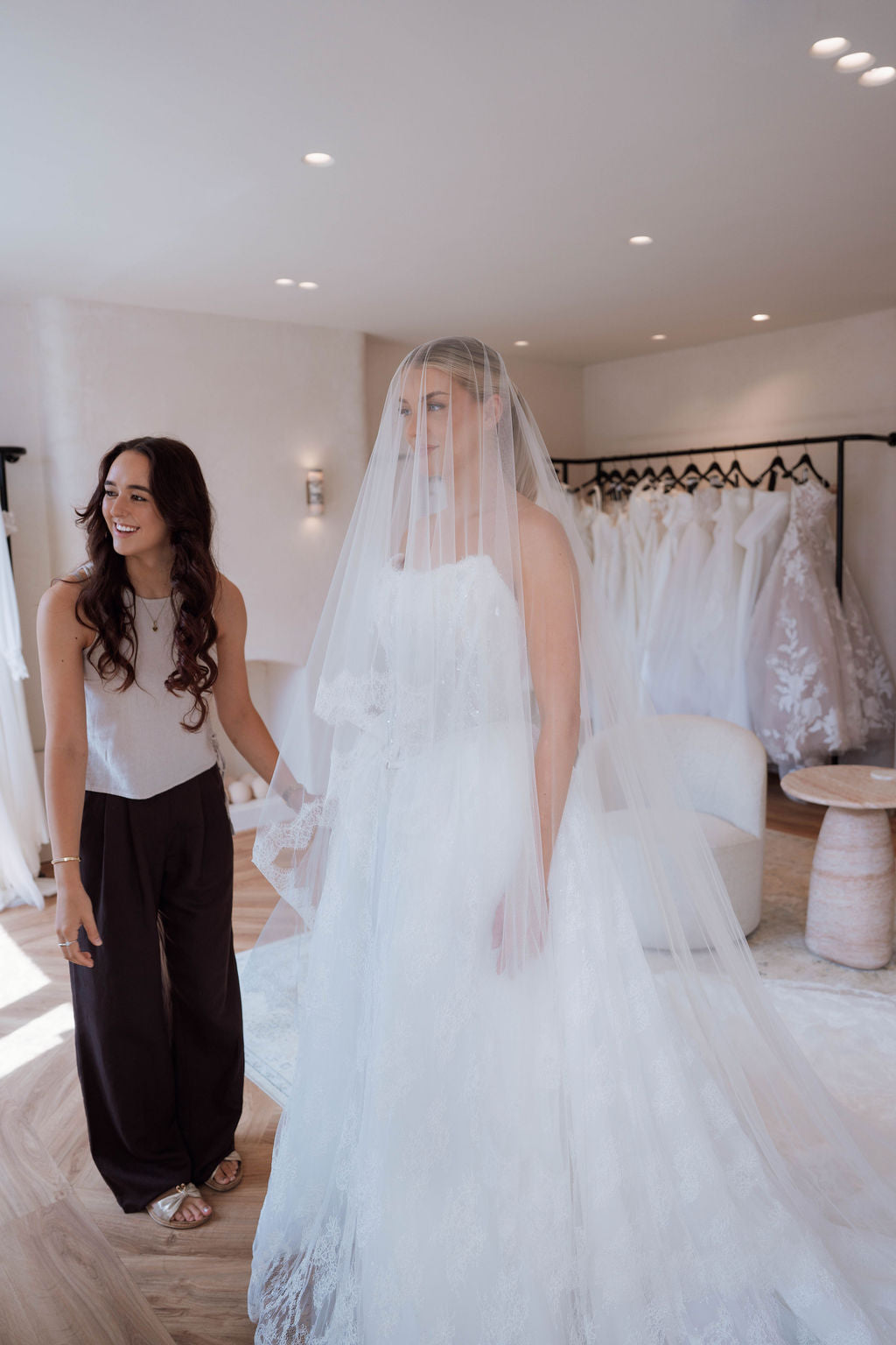 A bride in a white wedding gown and long veil walks through a bridal boutique, holding hands with a smiling woman in a white top and dark wide-leg pants. Wedding dresses are visible on a rack in the background.